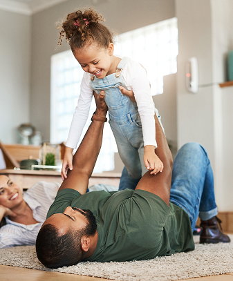 Man holding his daughter in his arms with his wife standing beside him, all smiling and enjoying a family moment