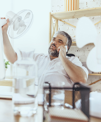 Man talking on the phone while holding an electric fan in his other hand, looking warm or overheated