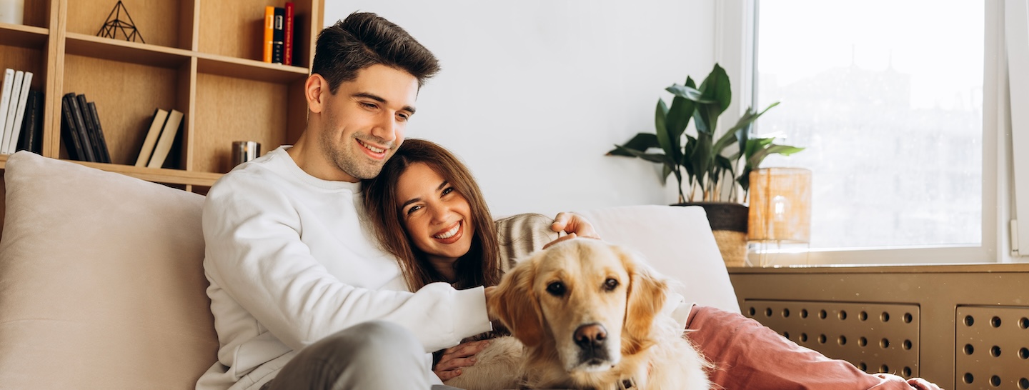 Young couple embracing and smiling while sitting on a comfortable sofa with their adorable golden retriever dog in a cozy living room