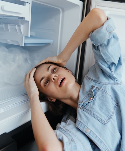 Young woman feeling overwhelmed, holding her head in front of an open refrigerator, expressing stress and frustration in a modern kitchen setting